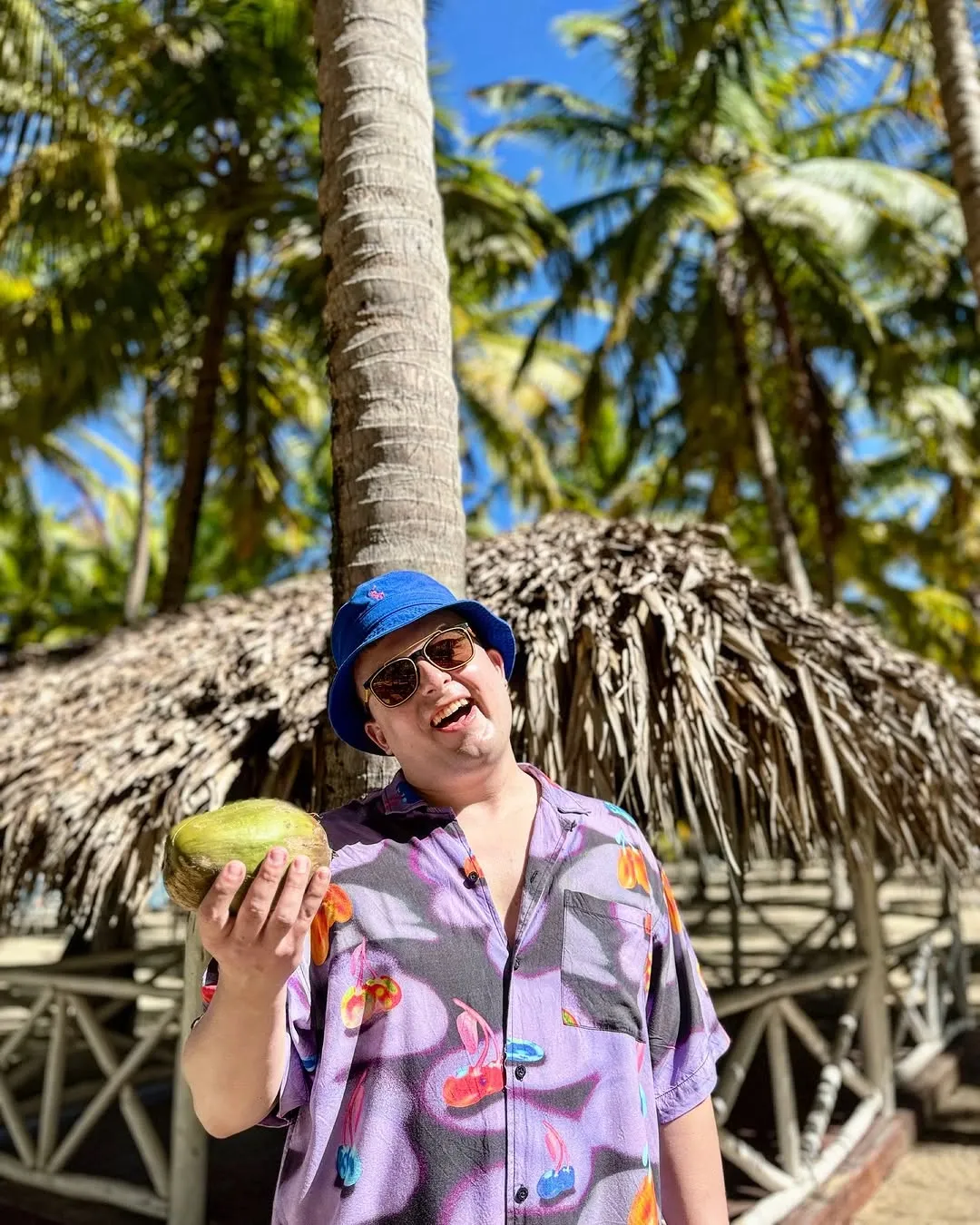A smiling man in a tropical shirt holding a fresh coconut under the palms